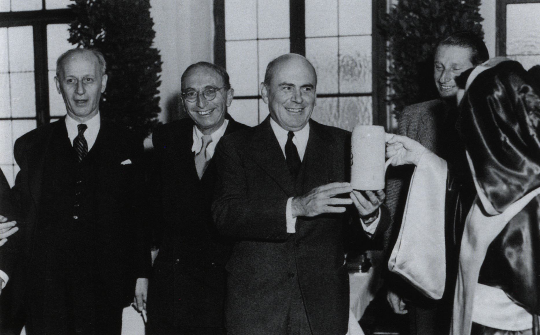 Der Landeskommissar George N. Shuster (ganz links) gemeinsam mit dem Hohen Kommissar John J. McCloy (Mitte) auf einer Veranstaltung in München; der <span class="highlight">High Commissioner</span> McCloy erhält aus der Hand des „Münchner Kindls“ (ganz rechts) eine Maß Bier. Aufnahme unbekannten Datums.<br/>Mit der Berufung des deutschstämmigen Hochschullehrers, Pädagogen und Publizisten George N. Shuster traten die Beziehungen zwischen der Staatsregierung und der Besatzungsmacht in eine neue Phase, nachdem das Verhältnis zu Shusters Vorgänger Clarence M. Bolds von starken Konflikten und Spannungen geprägt gewesen war. Nachdem die Amerikaner nach Inkrafttreten des Besatzungsstatuts gegenüber den deutschen Stellen ohnehin in zunehmendem Maße eine Politik der Nichteinmischung praktizierten, waren es nicht zuletzt auch die Persönlichkeit und die zurückhaltende Amtsführung des Katholiken und Deutschlandexperten George N. Shuster, die wieder für eine Normalisierung und atmosphärische Verbesserung der Beziehungen zwischen Staatsregierung und Landeskommissariat sorgten. Zusätzlich hatte das Landeskommissariat nach Gründung der Bundesrepublik und der Errichtung der Alliierten Hohen Kommission auf dem Petersberg bei Bonn auch merklich an Bedeutung verloren. Vor allem unter dem Vorzeichen der ab 1950 rapide anwachsenden legislativen Tätigkeit des Bundes suchte Ministerpräsident Ehard verstärkt den direkten persönlichen Kontakt zu McCloy, um bei einzelnen Gesetzgebungsverfahren die föderalen Interessen Bayerns zu wahren. Die von Bundeskanzler Adenauer 1950/51 geplante Errichtung einer Bundesbereitschaftspolizei beispielsweise kam aufgrund der Intervention Ehards bei McCloy nicht in der von Adenauer angestrebten Form zustande.<br/>University of Notre Dame Archives/Shuster Papers