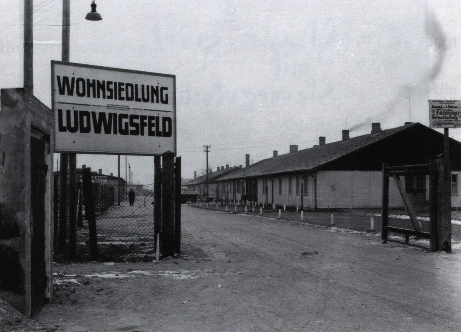 Der Eingang des ursprünglich von der Staatlichen Erfassungs-Gesellschaft für Öffentliches Gut m.b.H. (StEG) betriebenen Flüchtlingslager München-Ludwigsfeld. Im Oktober und November 1950 mußte die Bayerische Staatsregierung aufgrund des im September in New York gefaßten Beschlusses der Westalliierten, ihre Truppenpräsenz in Europa zu verstärken, innerhalb weniger Wochen eine Reihe von Kasernenanlagen räumen, die bis dahin als Flüchtlingslager genutzt wurden. Die so notwendig gewordene anderweitige Unterbringung der Flüchtlinge stellte die Staatsregierung vor große Probleme. Die Kasernenbewohner wurden z.T. auf andere, bereits bestehende Flüchtlingslager verteilt. Das hier abgebildete frühere StEG-Lager Ludwigsfeld, das im Jahre 1950 leer stand, wurde für die Aufnahme der umzusiedelnden Flüchtlinge durch die Landesflüchtlingsverwaltung ausgebaut.</br>Nachweis: Bayerisches Hauptstaatsarchiv, Abt. V, Bildersammlung 1326