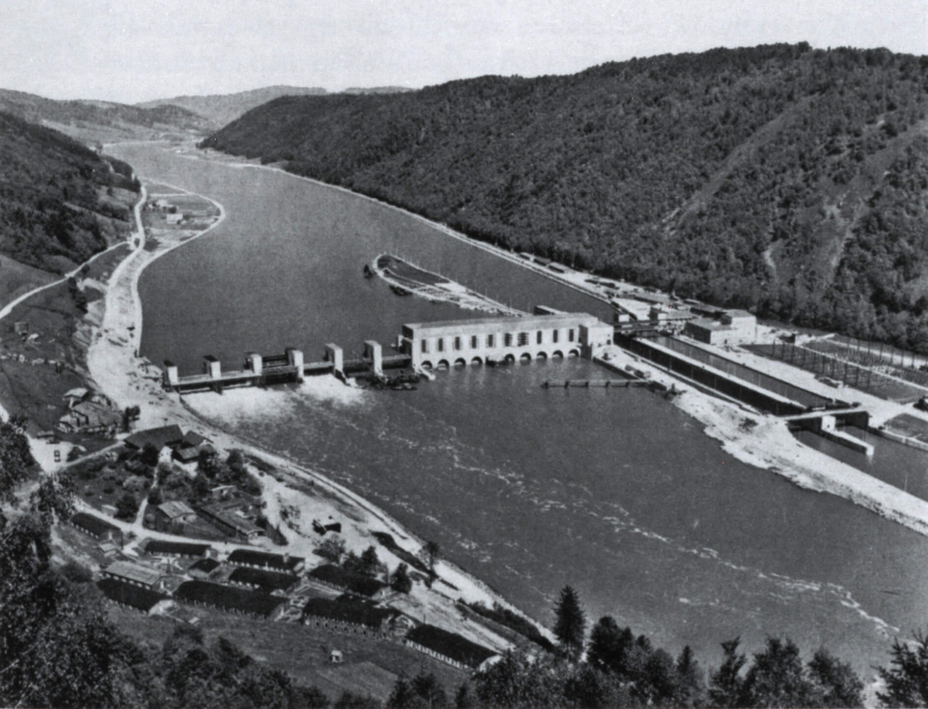 Luftaufnahme des Donaukraftwerks Jochenstein nach der vollständigen Inbetriebnahme mit fünf Turbinensätzen am 20. August 1956. Das Kraftwerk, dessen Bau Ende 1952 begonnen wurde und das zunächst ab Frühjahr 1955 nur mit drei Turbinenanlagen in Betrieb genommen wurde, war als deutsch-österreichisches Gemeinschaftsprojekt über der mittig in der Donau verlaufenden Landesgrenze zwischen den Gemeindegebieten des niederbayerischen Untergriesbach (rechts im Bild) und des oberösterreichischen Engelhartszell (links im Bild) errichtet worden<br/>Nachweis: Bayerisches Wirtschaftsarchiv, S 12/1031.