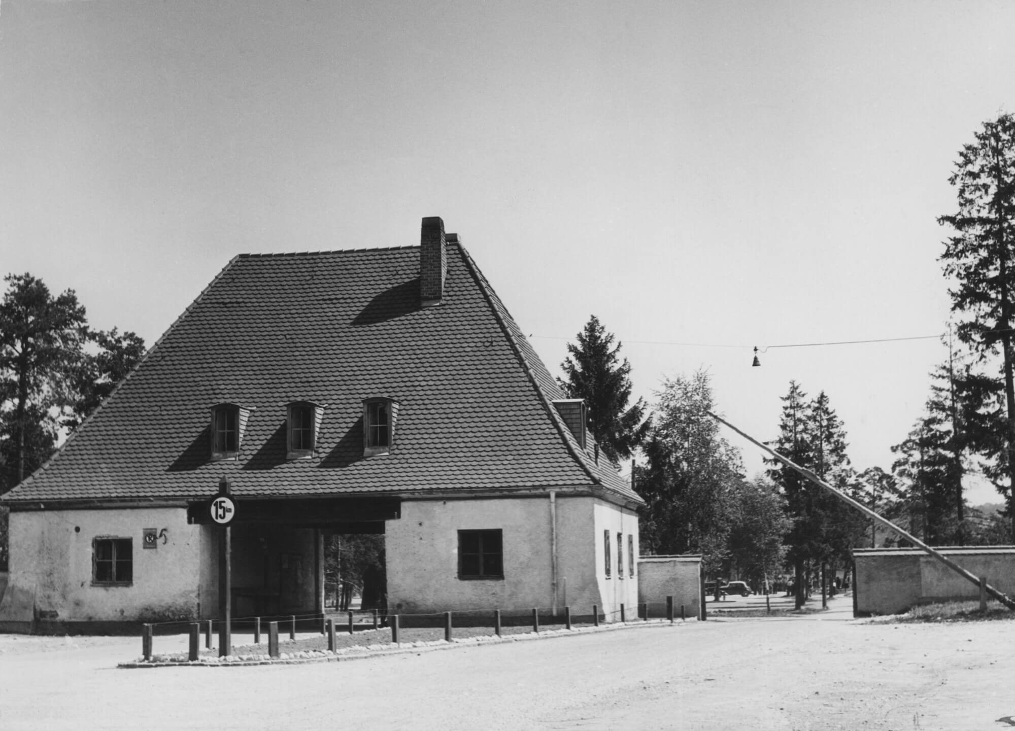 Zufahrt zum DP-Lager Föhrenwald bei Wolfratshausen. Ursprüngloch im Jahre 1939 erbaut als Wohnsiedlung für Arbeiter und Zwangsarbeiter zweier Munitionsfabriken, war Föhrenwald unmittelbar nach Kriegsende von der US-Besatzungsmacht zu einem internationalen DP-Lager, dann im September 1945 zu einem jüdischen DP-Lager erklärt worden. Pläne, das seit Dezember 1951 unter der Bezeichnung "Regierungslager für heimatlose Ausländer" in deutsche Verwaltung stehende Lager baldmöglichst aufzulösen, konnten nicht umgesetzt werden. 1952 befanden sich immer noch rung 2 000 Bewohner im Lager, diese Zahl erhöhte sich im Jahre 1953 noch durch die sogenannten "Illegalen". Die Frage der Behandlung dieser Remigranten, die aus Isreal über europäische Drittstaaten wieder in die Bundesrepublick zurückgekehrt waren, beschäftigte den Ministerrat wiederholt. Das Regierungslager Föhrenwald wurde erst Ende Februar 1957endgültig geschlossen.<br/>Nachweis: Stadtarchiv München FS-ER-C-0061.