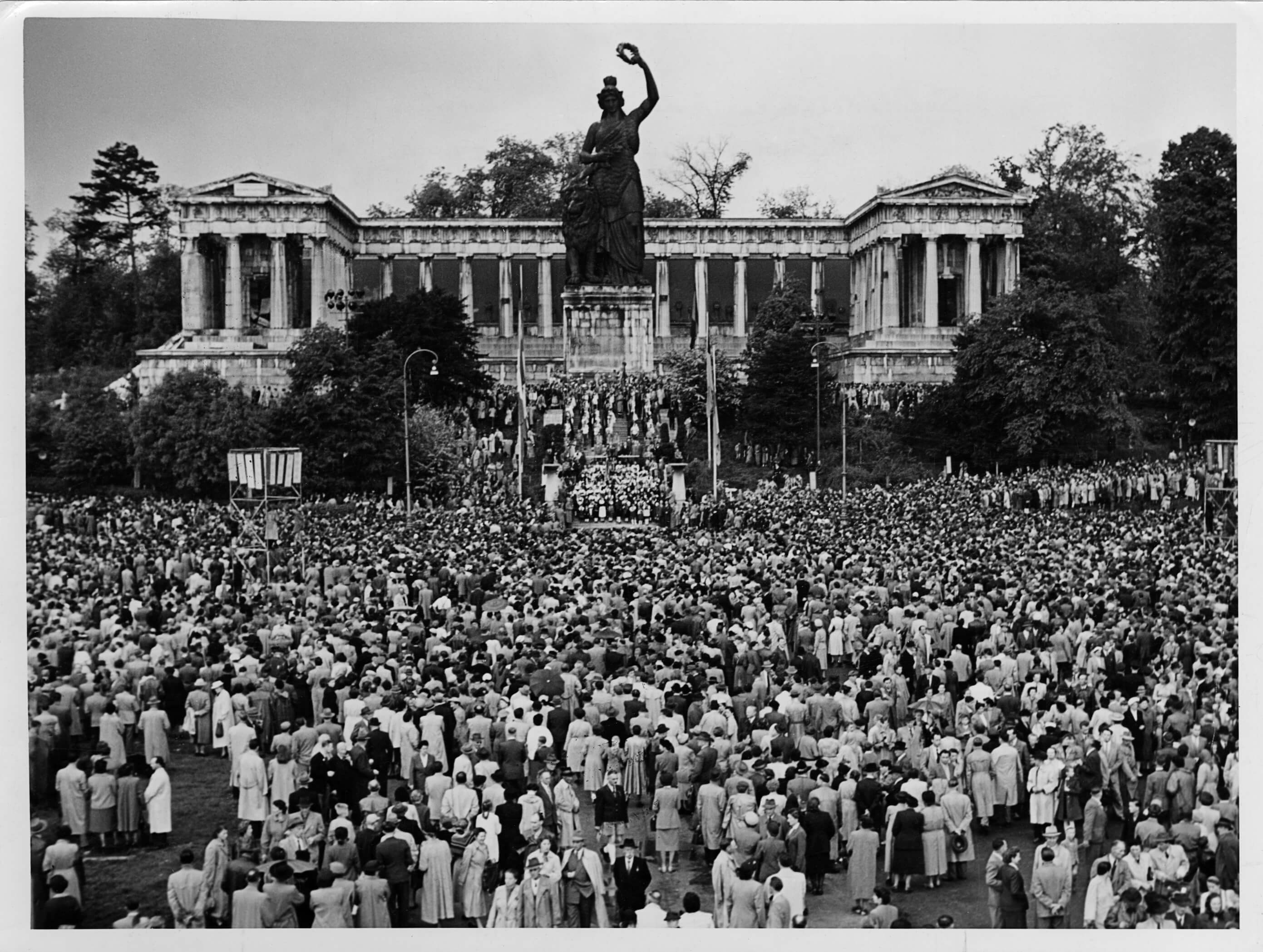 Versammlung von Teilnehmern einer Kundgebung auf dem Sudetendeutschen Tag 1954 vor der Bavaria auf der Münchner Theresienwiese. Als Mischung von Brauchtumsveranstaltung und politischer Großkundgebung zogen die seit 1950 jährlich an Pfingsten unter der Regie der Sudetendeutschen Landsmannschaft (SL) organisierten Sudetendeutschen Tage regelmäßig zehntausende, z.T. bis zu hunderttausende Teilnehmer an. Der Sudetendeutsche Tag in München vom 4. bis 7. Juni 1954 zählte an die 500.000 Besucher. Auf dem 5. Sudetentag in München verkündete Ministerpräsident Hans Ehard am 4. Juni 1954 die Schirmherrschaft des Freistaates über die Sudetendeutschen und sandte damit im Vorfeld der Landtagswahl 1954 ein wichtiges Signal für die politische und gesellschaftliche Anerkennung der Vertriebenen in Bayern. Auch wurden die Vertriebenen neben den Altbayern, den Franken und den Schwaben zum „Vierter Stamm“ Bayerns erhoben. Als im Ministerrat vom 18. Mai 1954 die Frage eines staatlichen Zuschusses zum Sudetendeutschen Tag 1954 erörtert wurde, äußerte Staatssekretär Stain noch die Fehleinschätzung, daß dieser „heuer zum letztenmal stattfinden werde, da seine Ziele im wesentlichen erreicht seien.“ Die Sudetendeutschen Tage finden bis in die Gegenwart weiterhin statt.<br/>Nachweis: SdA Bildersammlung 22339