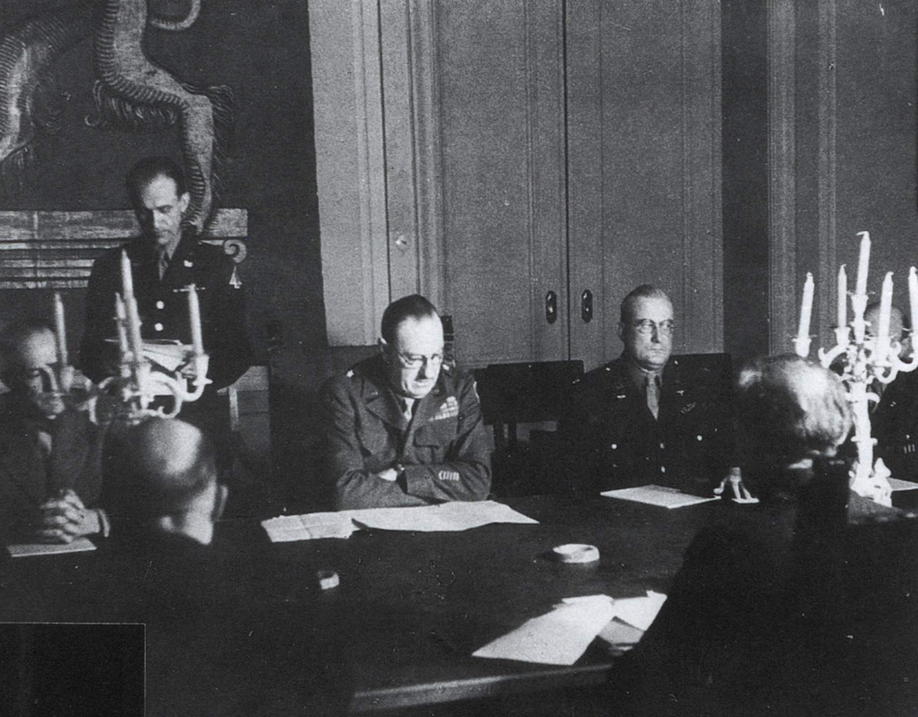 Das Photo zeigt eine Konferenz zwischen dem Direktor des Office of Military Government for Bavaria (OMGB) Brigadegeneral Walter J. Muller (1895–1967) und Abteilungsleitern von OMGB mit Ministerpräsident Wilhelm Hoegner und Mitgliedern seines Kabinetts. Ein Datum war nicht zu ermitteln. Ort der Sitzung war wahrscheinlich, dem Fuß des Bayerischen Löwen auf dem Wandteppich nach zu schließen, der Sitzungssaal des Bayerischen Wirtschaftsministeriums in der Prinzregentenstraße 28 (vgl. Kock, Bayerischer Landtag S. 37). Nur für eine Sitzung in dieser personellen Zusammensetzung, vom 21. März 1946, liegt ein Protokoll vor, das als Anhang zu den Ministerratsprotokollen abgedruckt wird. Sie fand, wie einige weitere, in der Bibliothek des Hauptquartiers von OMGB in der Tegernseer Landstraße 210 (Gebäude der Reichszeugmeisterei) statt.<br/>Privatbesitz Harald Hoegner, München